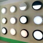 Round LED bulkhead wall lights in various designs and finishes, including half-moon cut, full diffuser, and black, white, and stainless-steel housings, displayed on showroom wall