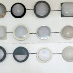Round and square LED bulkhead wall lights in various trims and finishes, including full-diffuser, half-moon, stainless steel, black, grey, and white housings, displayed on showroom wall